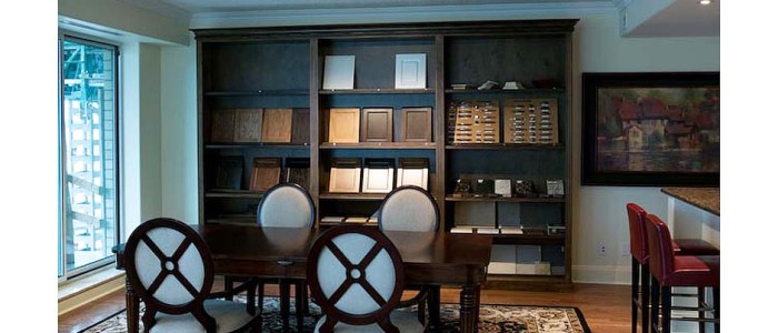 Condominium sample selection room with dark oak wall-unit in background holding sample selections, traditional wood dining table with 4 dark wood chairs white upholstered seats and backs in foreground sitting on oriental area rug