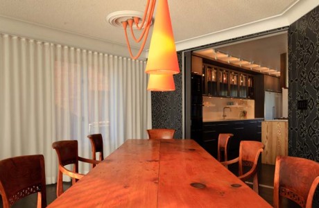 looking down the length of pine harvest dining table with orange pendant lights. walls in black and gray damask, sheer drapes on all exterior walls, kitchen beyond seen through large pocket doors
