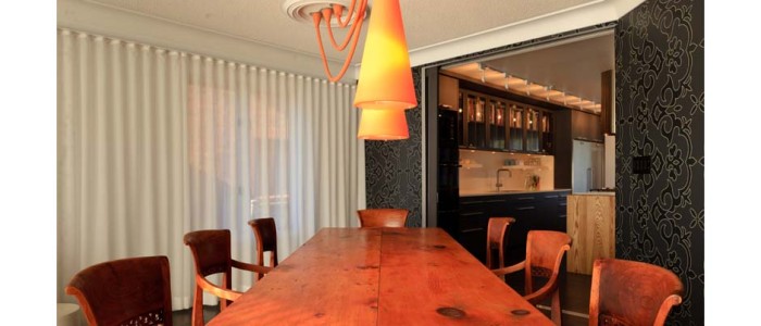 looking down the length of pine harvest dining table with orange pendant lights. walls in black and gray damask, sheer drapes on all exterior walls, kitchen beyond seen through large pocket doors