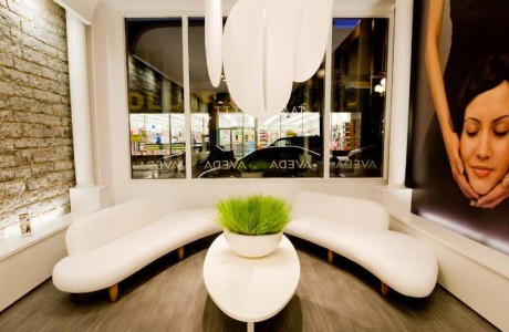 Modern hair salon waiting area with white semi circular leather sofas flanking white oval coffee table. large image on side wall of hands holding woman's head under chin