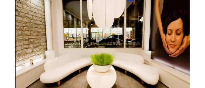 Modern hair salon waiting area with white semi circular leather sofas flanking white oval coffee table. large image on side wall of hands holding woman's head under chin