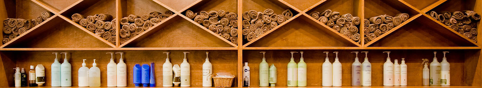 Open wall cabinet in wood. Lower shelf containing shampoo bottles, upper diamond shaped openings with rolled brown towels