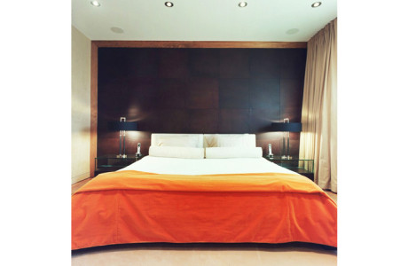 modern bed with orange comforter folded across the end, matching night tables and lamps on each side with chocolate brown leather tiles on wall behind