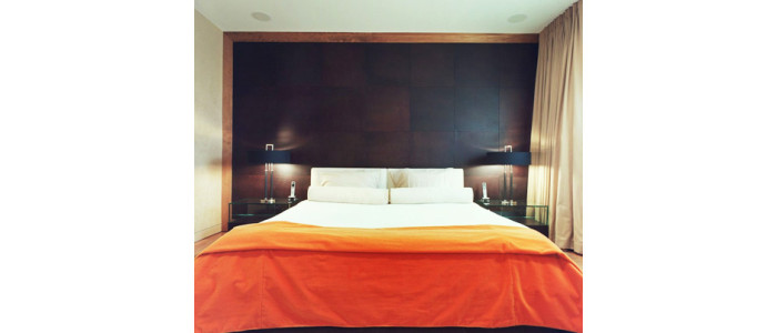 modern bed with orange comforter folded across the end, matching night tables and lamps on each side with chocolate brown leather tiles on wall behind