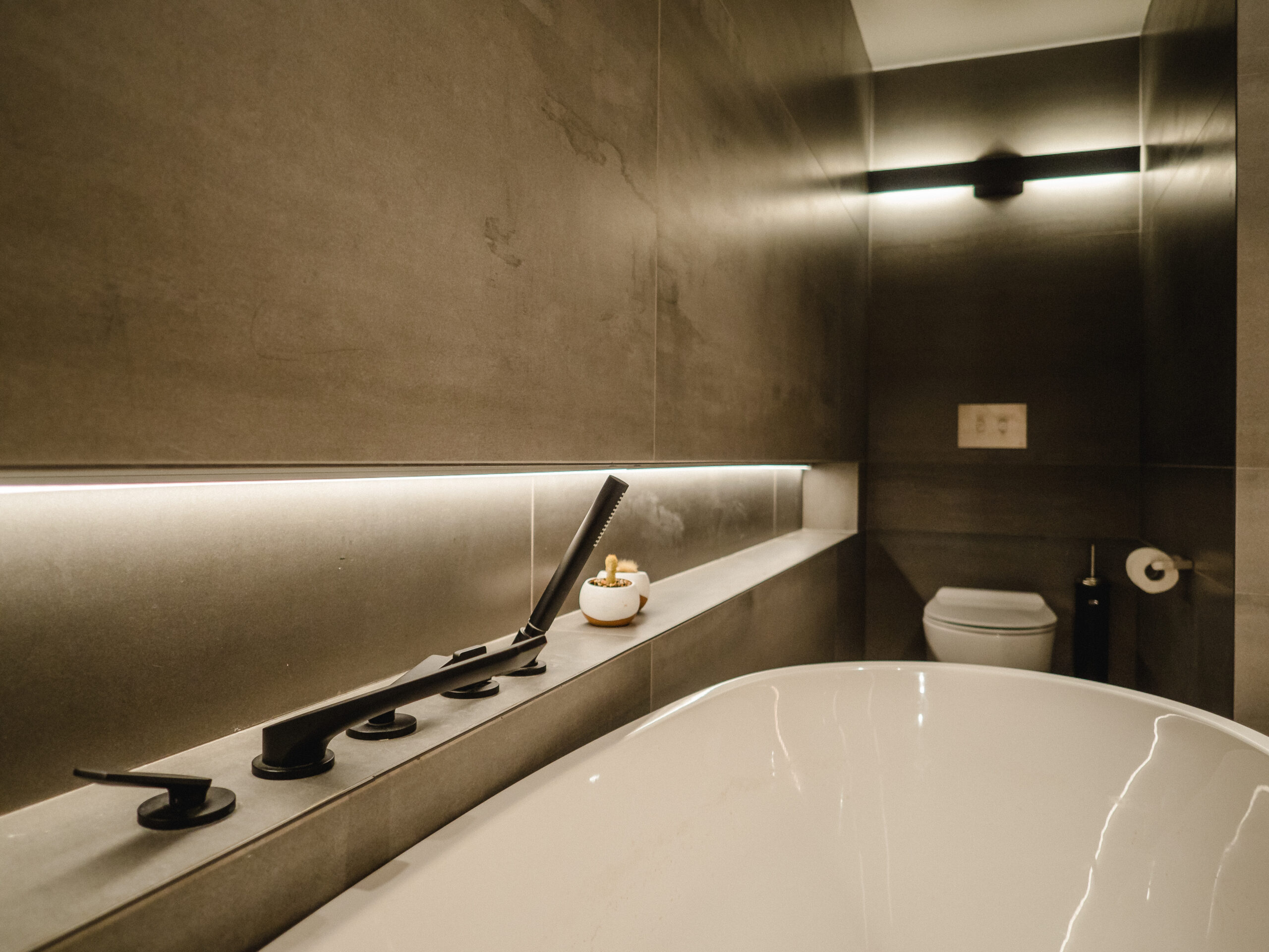 Freestanding white tub set against a dark grey tiled wall with built-in shelf, matte black tub fixtures, and a modern wall-mounted toilet with ambient lighting.