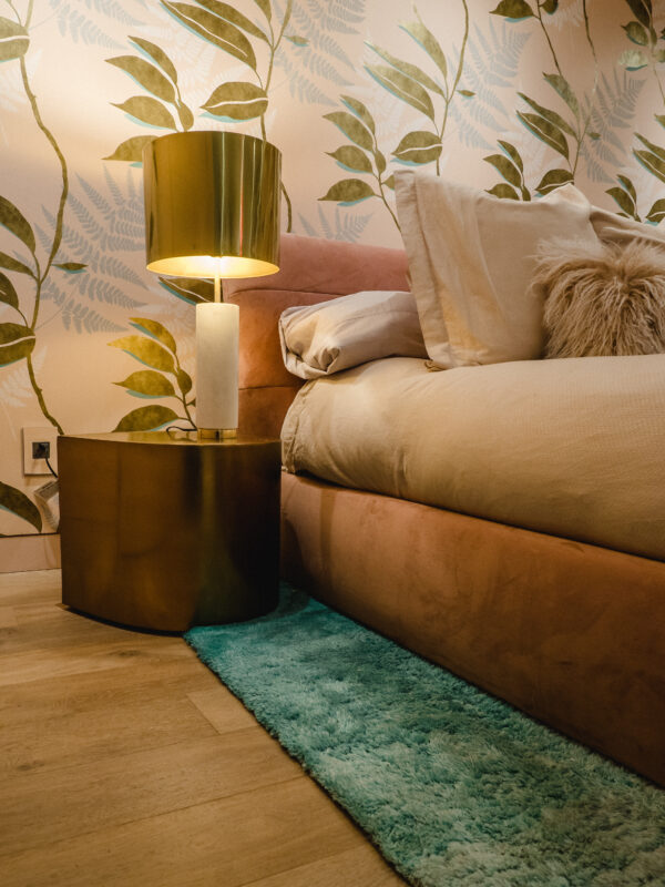 Detail of a cozy bedroom corner featuring a blush velvet bed, green botanical wallpaper, brass and marble lamp, and teal shag rug on hardwood flooring.