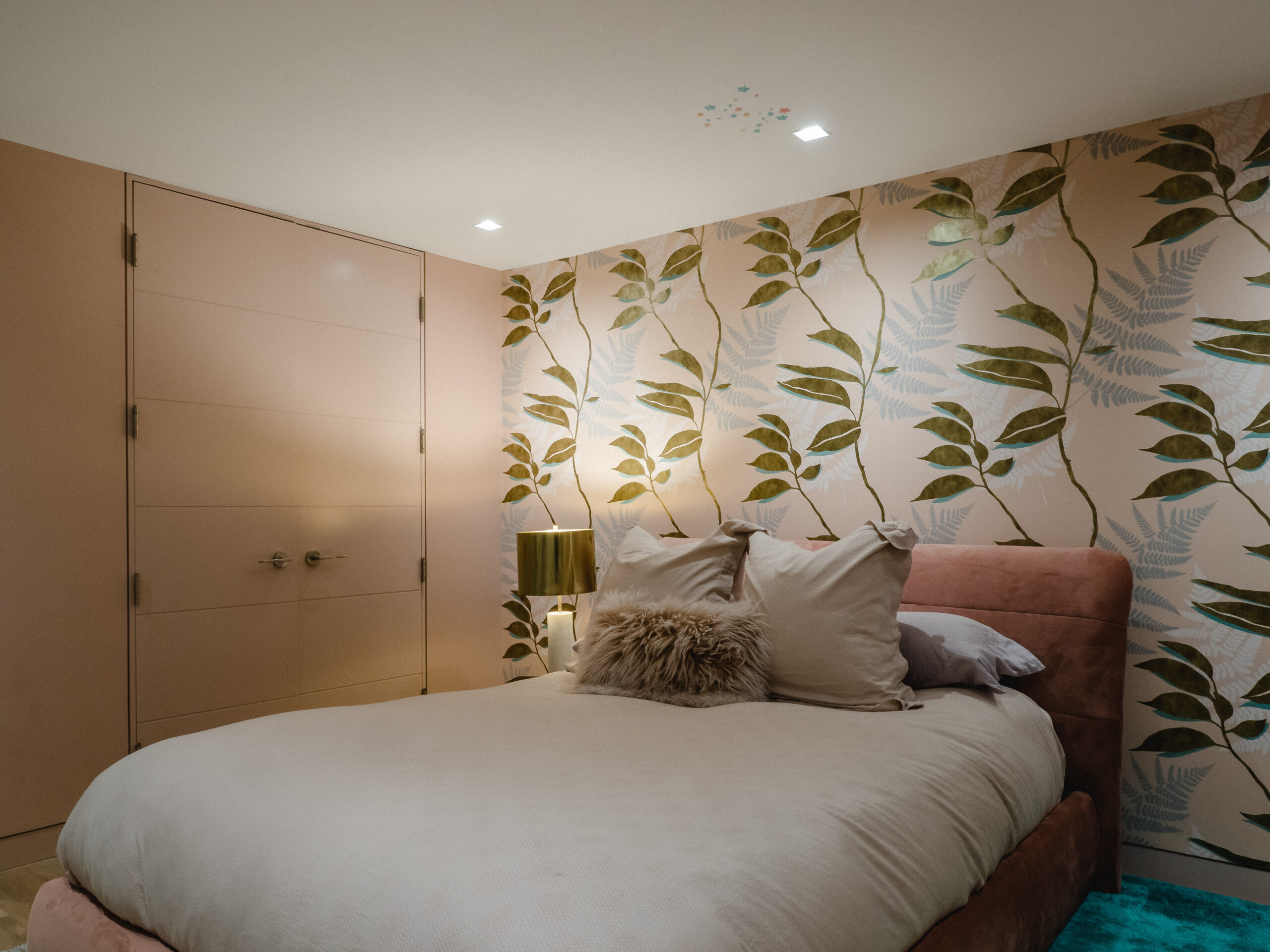 Playful basement bedroom with a blush-pink velvet bed, leafy botanical wallpaper, and pink-painted double doors adding charm and character.