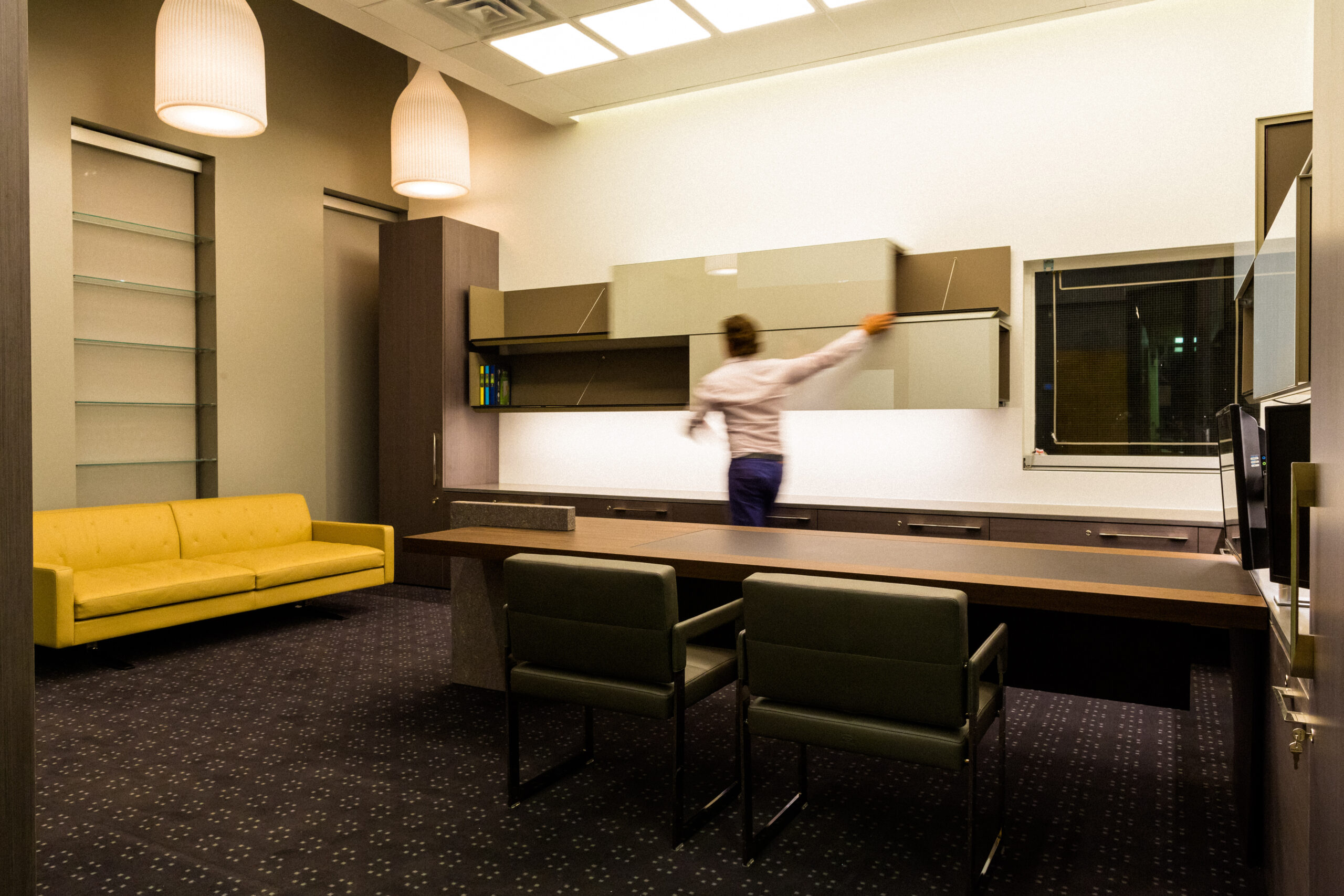 Modern executive office with custom wood desk, floating glass cabinets, integrated storage, and yellow sofa under sculptural pendant lights