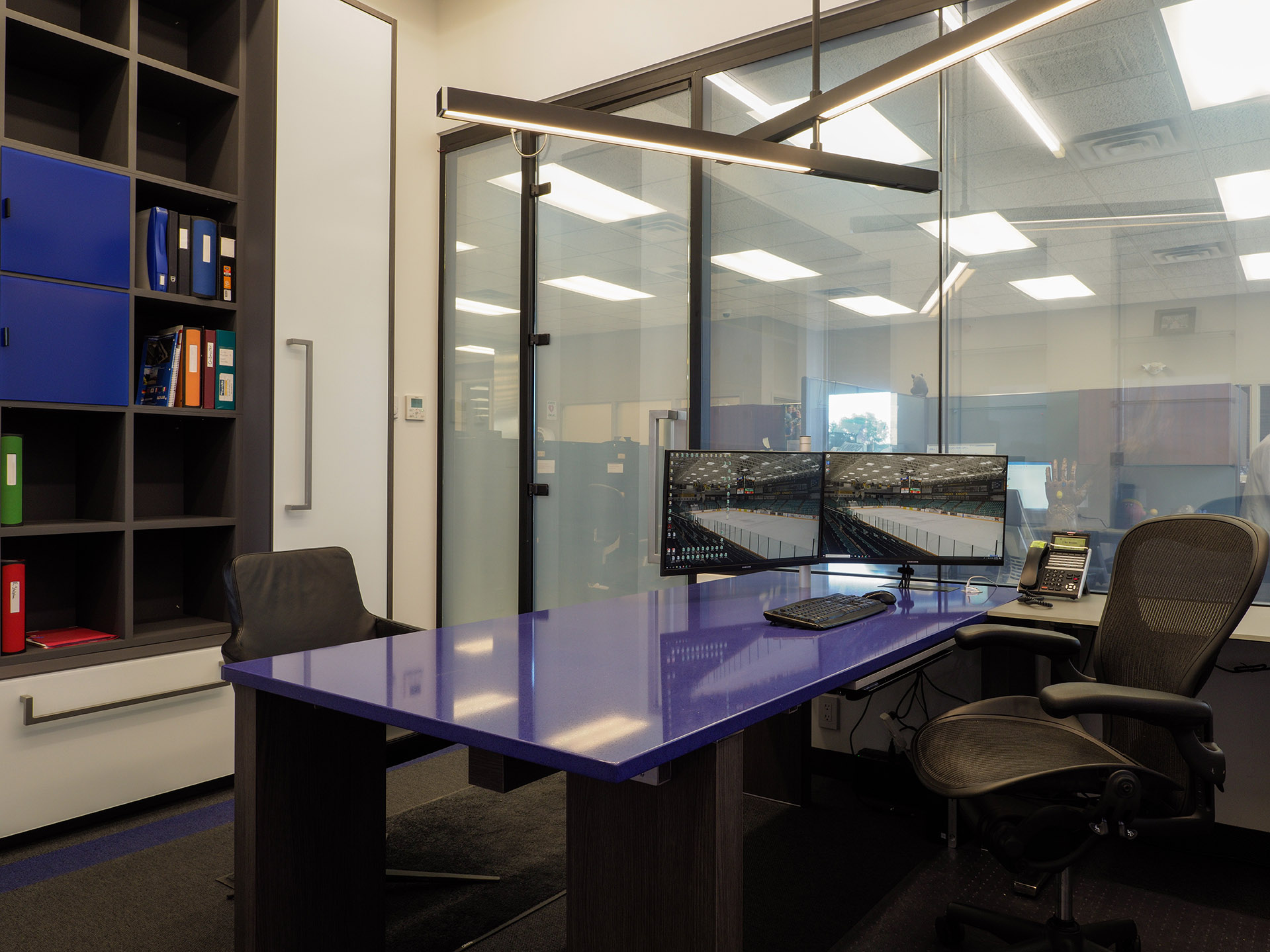 Executive workspace with dual monitors on a vivid blue quartz desk, set against a backdrop of custom millwork and full-height glass walls.