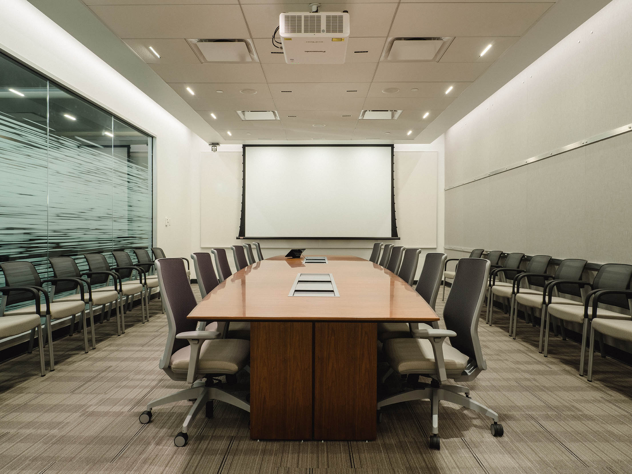 Contemporary boardroom design with glass walls, built-in AV system, and tiered guest seating along the perimeter.