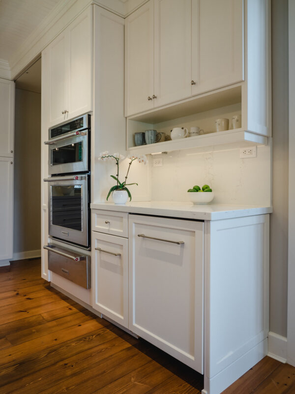 Detail of white shaker-style kitchen cabinetry with integrated appliances and an open shelf displaying dishware, finished with a clean quartz backsplash and natural wood floors.