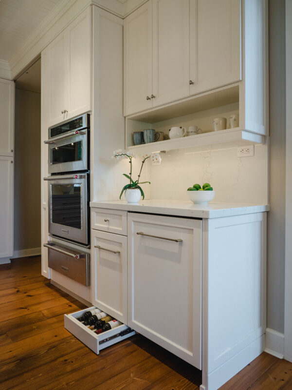 Custom white kitchen cabinetry with a hidden pull-out wine drawer, built-in double wall ovens, and display shelf with neutral ceramic mugs, set against a backdrop of warm wood flooring.